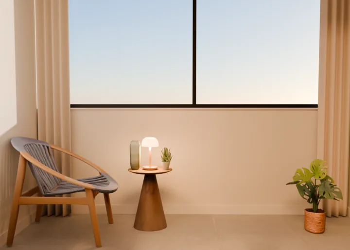 Varanda envidraçada no fim da tarde, vista da sala: poltrona de madeira com assento de corda azul à esquerda, mesa lateral cônica com a luminária acesa, um vaso verde e uma suculenta, costela-de-adão num vaso de palha à direita, cortina de linho aberta nos dois cantos e o céu pela janela
